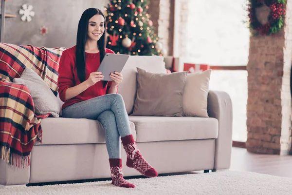 Фото милой юной леди сидеть диван держать планшет зубастый улыбающийся носить красный свитер джинсы джинсы носки в украшенной гостиной в помещении — стоковое фото