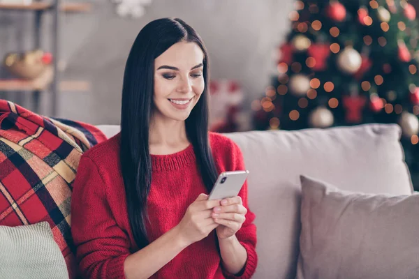 Fotografie krásné mladé dívky sedět pohovka držet smartphone lesklý úsměv nosit červený svetr v zdobeném obývacím pokoji uvnitř — Stock fotografie