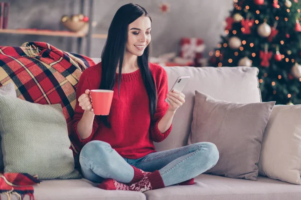 Фото молодой красивой девушки сидеть диван держать кружку телефон смотреть экран носить красные носки свитера джинсы в украшенной гостиной в помещении — стоковое фото