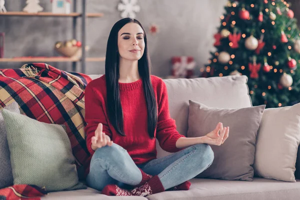 Fotografie mladé okouzlující žena sedět na gauči lotos pózovat oči zavřené nosit červené svetr džíny ponožky v zdobené obývací pokoj doma — Stock fotografie