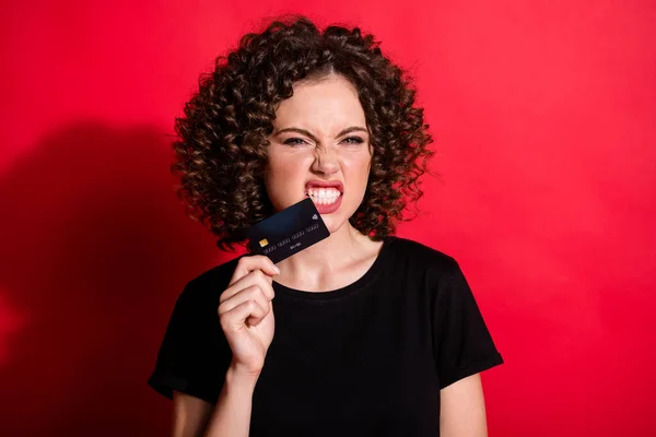 Foto de engraçado doce encaracolado mulher desgaste casual preto roupa segurando mordendo cartão de crédito isolado cor vermelha fundo — Fotografia de Stock