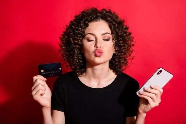 Foto av flörtig glänsande vågiga dam klädd casual svart outfit innehav kreditkort gadget läppar pip isolerad röd färg bakgrund — Stockfoto