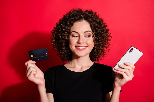 Foto av lockigt söt kvinna bära casual svart outfit innehav kreditkort ser mobiltelefon isolerad röd färg bakgrund — Stockfoto