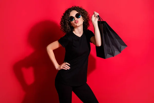 Foto van schattig schattig krullend vrouw dragen casual zwarte outfit bril holding tassen verzenden kus geïsoleerde rode kleur achtergrond — Stockfoto