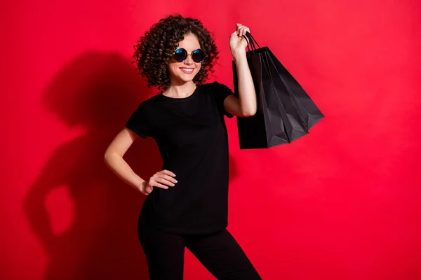 Foto van vrolijke glanzende golvende dame gekleed casual zwarte outfit brillen houden boodschappentassen geïsoleerd rode kleur achtergrond — Stockfoto