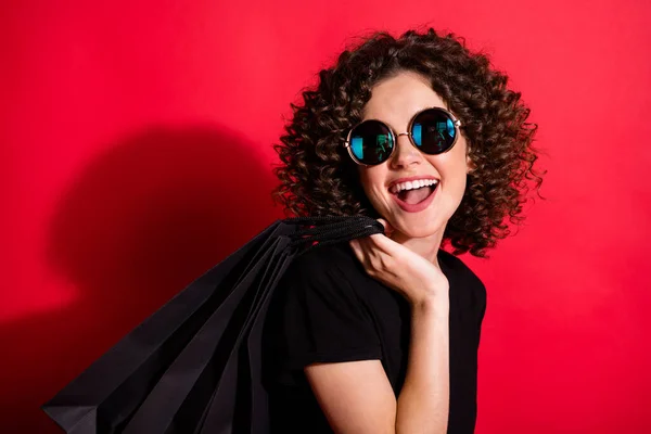 Foto av söt glad vågig dam klädd casual svart outfit glasögon hålla tillbaka shoppingväskor ser tomt utrymme isolerad röd färg bakgrund — Stockfoto