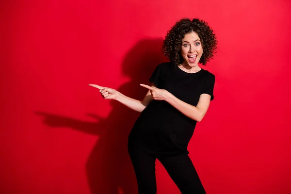 Foto de mujer rizada divertida sorprendida usar traje negro casual apuntando dos dedos espacio vacío aislado fondo de color rojo — Foto de Stock