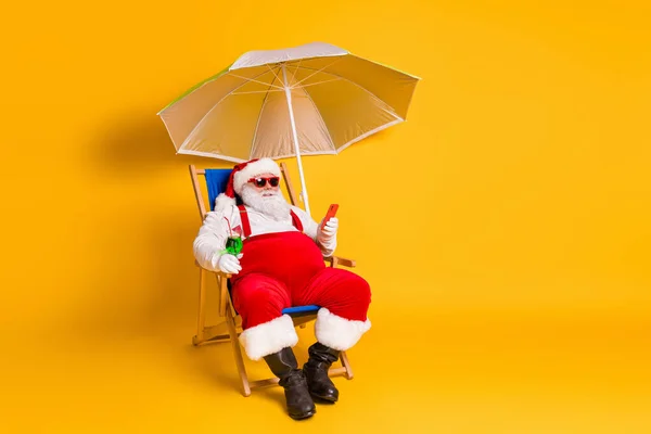 Pełny rozmiar zdjęcie siwych białych włosów brodaty Santa Claus chill chaise-lounge używać smartfona trzymać koktajl x-mas reszta nosić czerwony kapelusz kostium okulary przeciwsłoneczne odizolowany jasny połysk kolor tło — Zdjęcie stockowe
