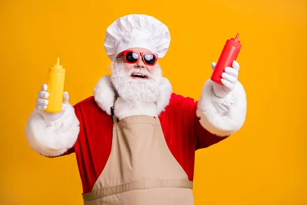 シェフの頭皮の白い灰色のひげの毛のサンタクラス料理のxマスクリスマスパーティースナックホールドマスタードトマトソースボトルはサングラスエプロン孤立した明るい輝きの色の背景を身に着けています — ストック写真