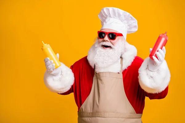 Photo of positive santa claus chef hold mustard tomato ketchup sauce bottle x-mas eve noel snack wear headwear sunglass apron isolated over yellow shine color background — Stock Photo, Image