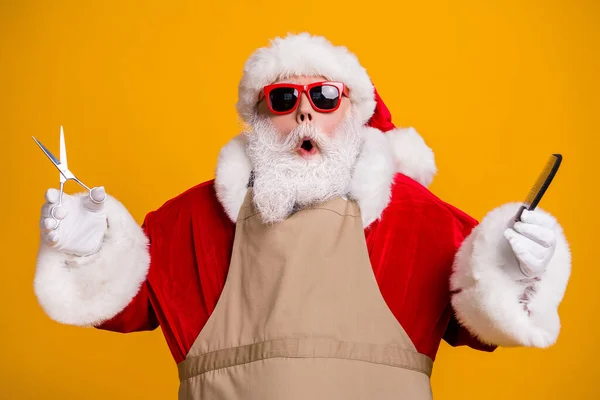 Close-up portrait of nice attractive stunned amazed funny grey-haired Santa wearing apron holding in hands coiffure equipment beauty salon isolated bright vivid shine vibrant yellow color background — Stock Photo, Image