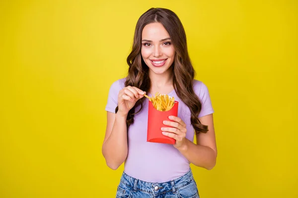 Porträtt av charmiga smala glada vågiga hår flicka äter pommes frites fritid isolerad ljus gul färg bakgrund — Stockfoto