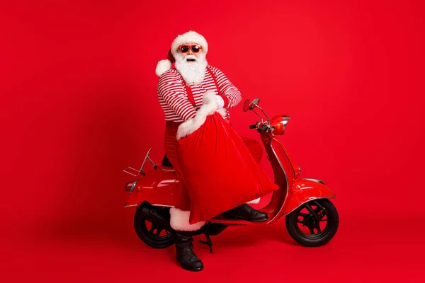 Pełna długość zdjęcie dziadka szara broda stojak w pobliżu retro motorower trzymać torba prezenty nosić Santa Claus x-mas kostium szelki sunglass paski koszula buty czapka odizolowany czerwony kolor tło — Zdjęcie stockowe