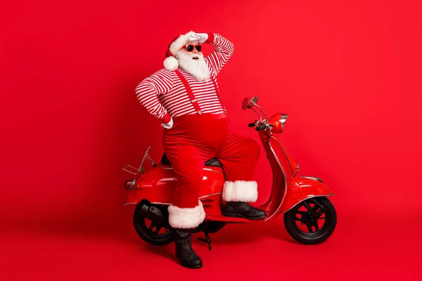 Volledige lengte foto van grootvader grijze baard staan in de buurt retro scooter kijken ver weg dragen santa claus x-mas kostuum bretels zonneglas gestreepte shirt laarzen cap geïsoleerde rode kleur achtergrond — Stockfoto