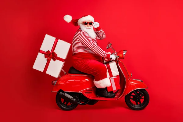 Volledige lengte profiel foto van opa grijze baard rijden retro brommer leveren doos slijtage kerstman x-mas kostuum bretels zonneglas gestreepte shirt cap laarzen geïsoleerde rode kleur achtergrond — Stockfoto