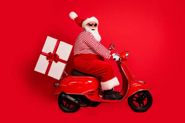 Dedemin tam boy profil fotoğrafı. Gri sakallı, retro scooter 'lı, hediye getiren Noel Baba, Noel Baba, Noel Baba, kostüm askısı güneş gözlüğü çizgili gömlek çizmeleri, izole edilmiş kırmızı arka plan. — Stok fotoğraf