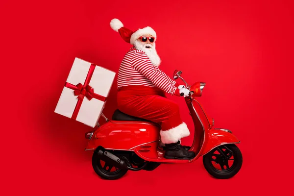 Volledige lengte profiel foto van grootvader grijze baard rijden retro scooter leveren gift wear kerstman x-mas kostuum bretels zonneglas gestreepte shirt laarzen cap geïsoleerde rode kleur achtergrond — Stockfoto