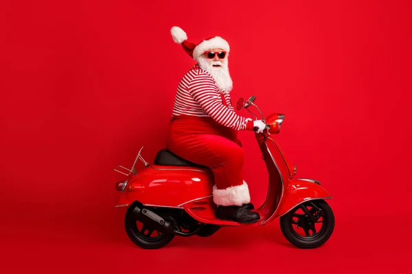 Volledige lengte profiel foto van opa grijze baard rijden retro scooter look camera dragen kerstman x-mas kostuum bretels zonneglas gestreepte shirt cap geïsoleerde rode kleur achtergrond — Stockfoto