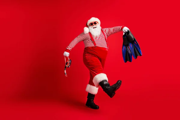 Pełna długość zdjęcie dziadka szara broda iść trzymać niebieski bieg maska płetwy nosić Santa Claus x-mas kostium szelki sunglass paski koszulka czapka buty odizolowany czerwony kolor tło — Zdjęcie stockowe