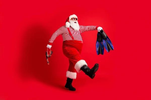 Full length profile photo of grandfather grey beard walk hold mask fins wear santa claus x-mas costume suspenders sunglass striped shirt hat boots isolated red color background — Stock Photo, Image