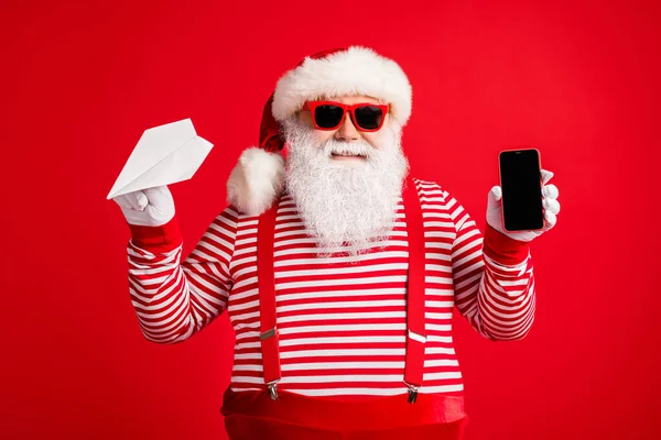 Foto van oude man grijze baard houden origami vliegtuig telefoon scherm lege ruimte dragen kerstman x-mas kostuum bretels zonneglas gestreepte shirt cap geïsoleerde rode kleur achtergrond — Stockfoto
