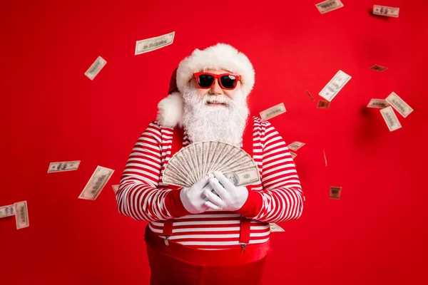 Foto di pensionato vecchio nonno tenere ventilatore contanti preparare spendere soldi newyear carità indossare bretelle costume di Babbo Natale camicia a righe unglass copricapi isolato colore rosso sfondo — Foto Stock