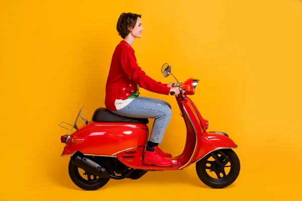 Volledige lengte profiel zijkant foto van positief meisje rijden rode scooter reizen x-mas tijd avond noel partij dragen kerst boom decor pullover denim jeans geïsoleerde heldere glans kleur achtergrond — Stockfoto