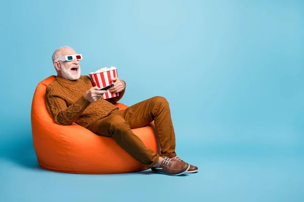 Çekici, neşeli, neşeli, sakallı, gri saçlı adamın portresi torba koltuğunda oturmuş mısır yerken eğleniyor mavi pastel arka planda izole edilmiş televizyon komedisi izliyor. — Stok fotoğraf