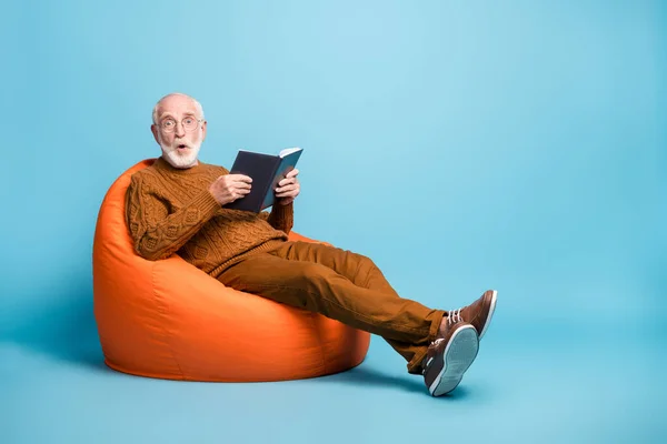 Güzel, çekici, şaşkın, sakallı, gri saçlı adamın portresi çanta koltuğunda oturuyor, kitap okuyor, dudak büküyor mavi pastel arka planda izole edilmiş yeni bilgiler ediniyor. — Stok fotoğraf