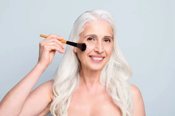 Foto de cerca de una hermosa mujer jubilada que aplica maquillaje de polvo de pómulo de cepillo fondo de color gris aislado — Foto de Stock