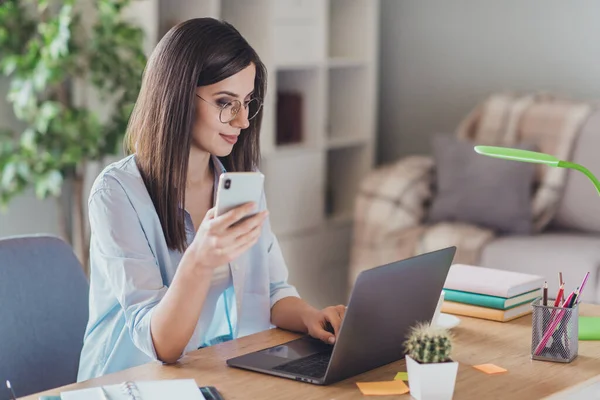 Foto de linda chica joven sostener teléfono inteligente mirada netbook pantalla usar gafas camisa en casa oficina en interiores —  Fotos de Stock
