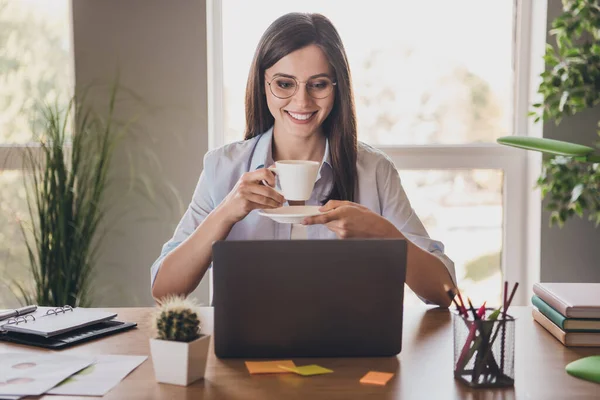 원문 기사보기 매력적 인 여성이 데스크 탑 팔에 앉아 컵 접시비친 미소가 실내 사무실에서 스펙 셔츠를 입고 있는 모습 — 스톡 사진