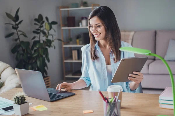 Foto von hübschen entzückenden Mädchen sitzen Tisch aussehen Bildschirm Computer halten Tablet tragen Brille Hemd im Home Office drinnen — Stockfoto