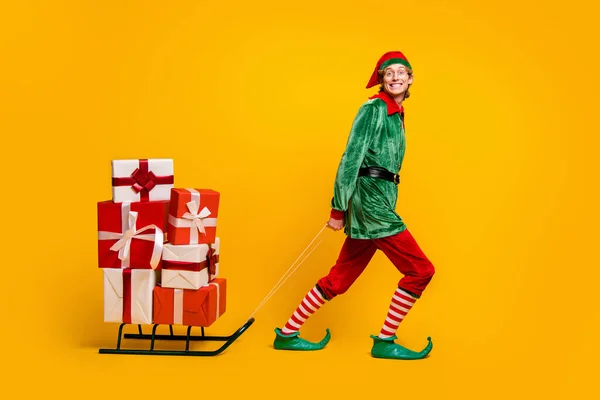 Pleine longueur taille du corps vue de son il agréable gai joyeux joyeux heureux gars drôle elfe portant de nombreuses boîtes-cadeaux sur luge fée isolé sur lumineux éclat vif fond de couleur jaune vibrant — Photo