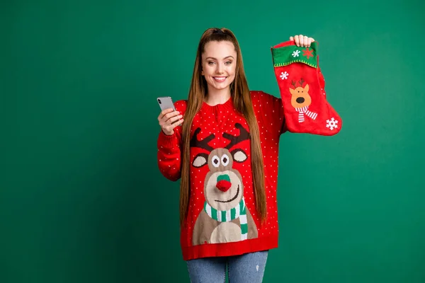 Photo of attractive lady hold large newyear stocking for x-mas eve present hold telephone shopping online internet buy gifts for family wear red ugly pullover isolated green color background — Stock Photo, Image