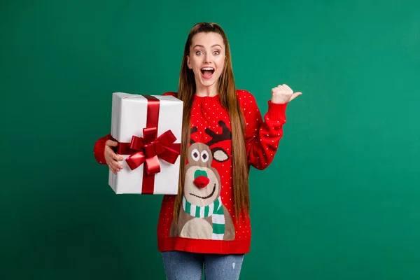 Фотографія привабливої шокованої леді тримає велику подарункову коробку новорічну подарункову коробку прямий палець збоку порожній простір x-mas продажу знижка на одяг червоний потворний пуловер ізольований зелений колір фону — стокове фото
