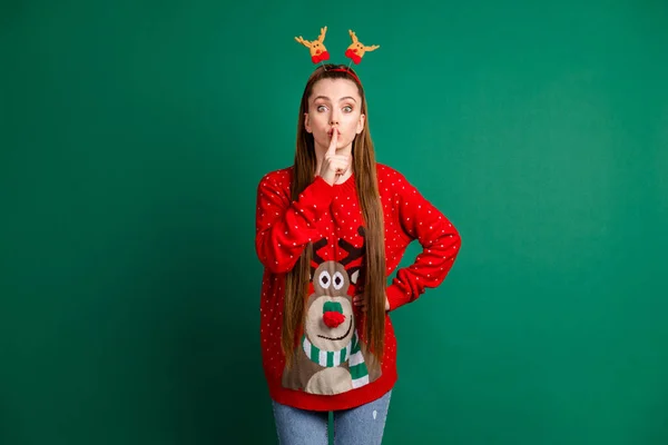 Photo of attractive cute lady long brown tail finger on lips ask friends keep silence boyfriend x-mas surprise wear deer horns headwear red ornament pullover isolated green color background — Stock Photo, Image