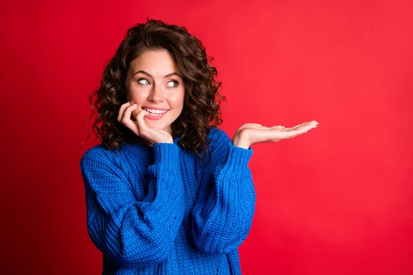 Photo of pretty charming lady curly hairstyle hand hold look empty space arm cheekbone cozy gnaw nail toothy smile wear blue knitted pullover isolated red color background — 스톡 사진