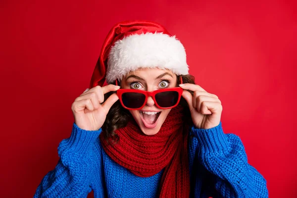 Foto ritratto di carina ragazza sorpresa togliersi tenendo gli occhiali con le dita bocca aperta indossando blu pullover sciarpa Santa cappello isolato su sfondo rosso vivido — Foto Stock