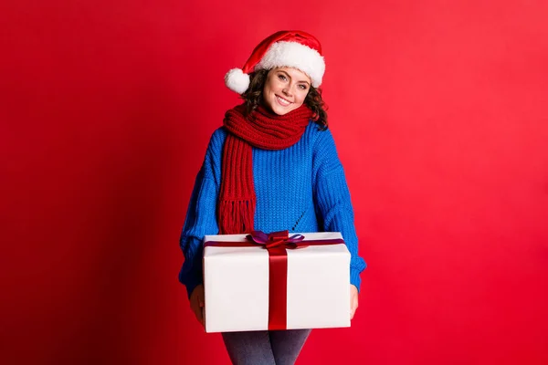 Fénykép portré bájos lány gazdaság jelen van elöl mosolygós visel santa kalap kék pulóver elszigetelt élénk piros színű háttér — Stock Fotó