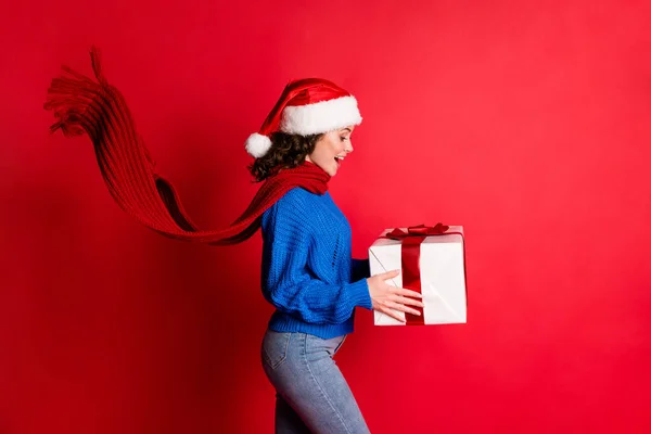 Foto retrato de hermosa chica excitada sosteniendo regalo usando bufanda de jersey azul fluyendo vista lateral del sombrero de santa aislado sobre fondo de color rojo vivo — Foto de Stock