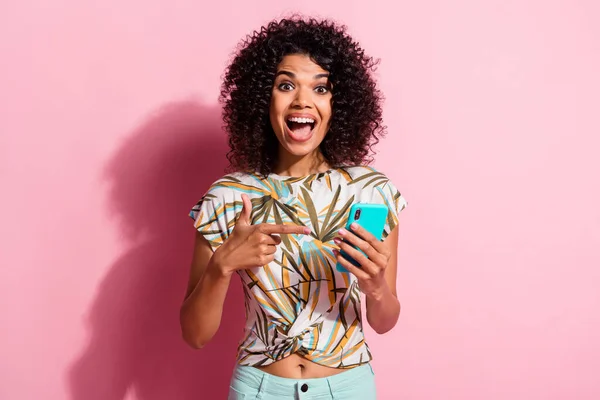 Retrato fotográfico de una mujer excitada con la boca abierta sosteniendo el teléfono en una mano señalándolo con el dedo aislado sobre un fondo de color rosa pastel — Foto de Stock