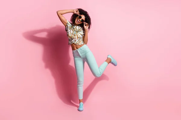 Foto de tamaño completo del cuerpo de la divertida chica de piel negra feliz bailando en el suelo tocando gafas de sol aisladas sobre fondo de color rosa pastel —  Fotos de Stock