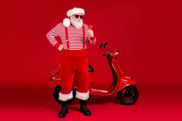 Comprimento total do corpo tamanho vista de sua ele bonito focado barbudo Papai Noel equitação ciclomotor usando gadget bate-papo post comentário feedback isolado brilhante vívido brilho vibrante cor vermelha fundo — Fotografia de Stock