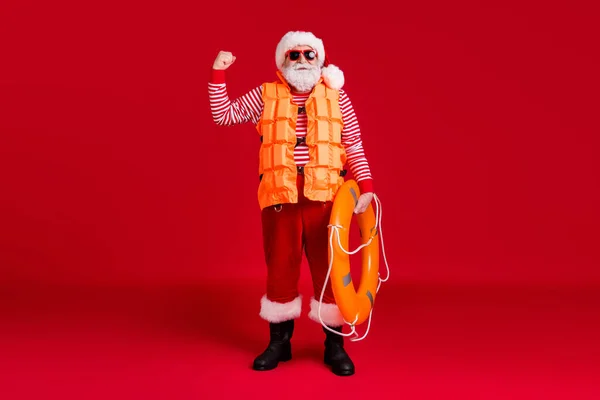 Pełna długość zdjęcie emerytowany szary broda stary człowiek trzymać nadmuchiwane koło podnieść ramię pokazać mięśnie nosić kamizelka Santa kostium szelki buty w paski koszula czapka odizolowany czerwony kolor tło — Zdjęcie stockowe