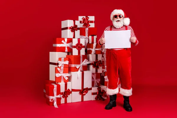 Volledige lengte foto van gepensioneerde opa grijze baard stack giftboxen houden bordje lege ruimte dragen santa kostuum bretels zonneglas laarzen gestreepte shirt broek cap geïsoleerde rode kleur achtergrond — Stockfoto