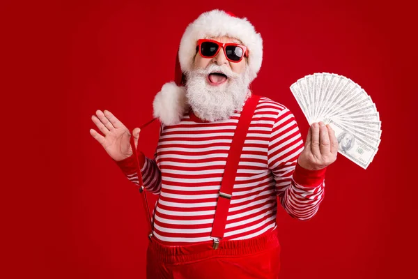 Zdjęcie emeryta stary człowiek szary broda trzymać pieniądze gotówka wentylator grać pasek zabawy milioner nosić Santa X-mas stroje szelki okulary w paski koszula czapka odizolowany czerwony kolor tło — Zdjęcie stockowe