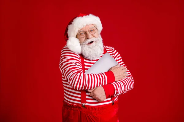 Portrait de son il beau beau gai joyeux père Noël barbu tenant dans les mains étreignant ordinateur portable pôle Nord Décembre hiver isolé lumineux vif éclat vibrant fond de couleur rouge — Photo
