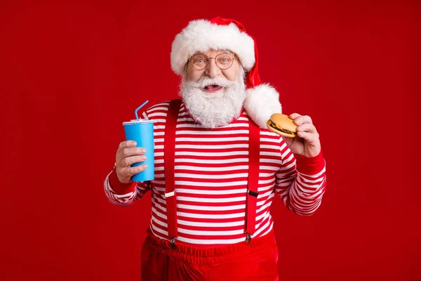 年金受給者の写真おじいちゃんグレーのひげホールドカップソーダバーガー小売リモートオーダーレストランは、サンタの衣装のサスペンダー眼鏡ストライプシャツの頭の摩耗隔離された赤の色の背景 — ストック写真