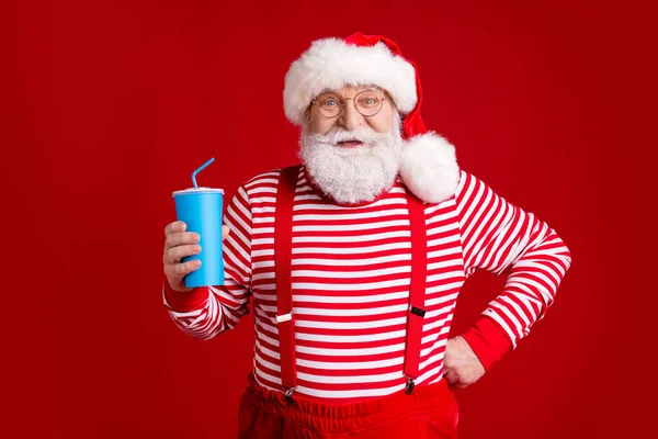 Photo of retired old man grey beard self-assured hold cup soda hand hip prepare quench thirst wear santa costume suspenders spectacles striped shirt headwear isolated red color background